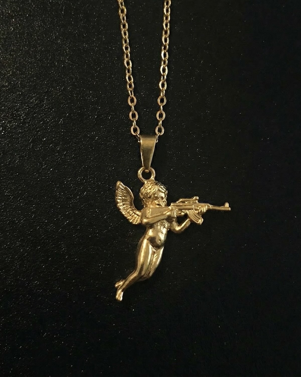 Gold cherub pendant shaped into a winged baby angel in flight holding a rifle, close detail